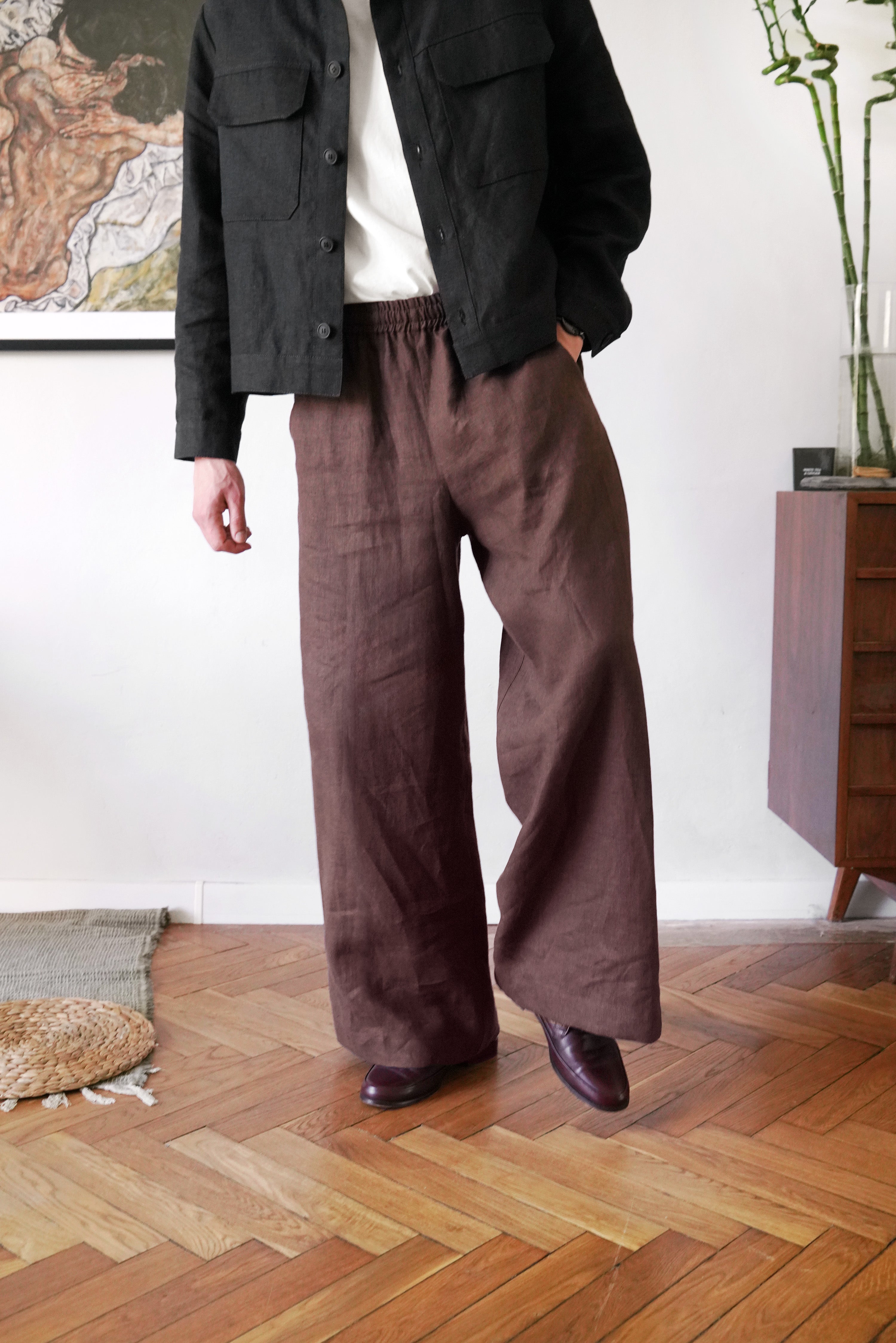 Person wearing a dark jacket and brown wide-leg pants standing in a room with wooden flooring.
