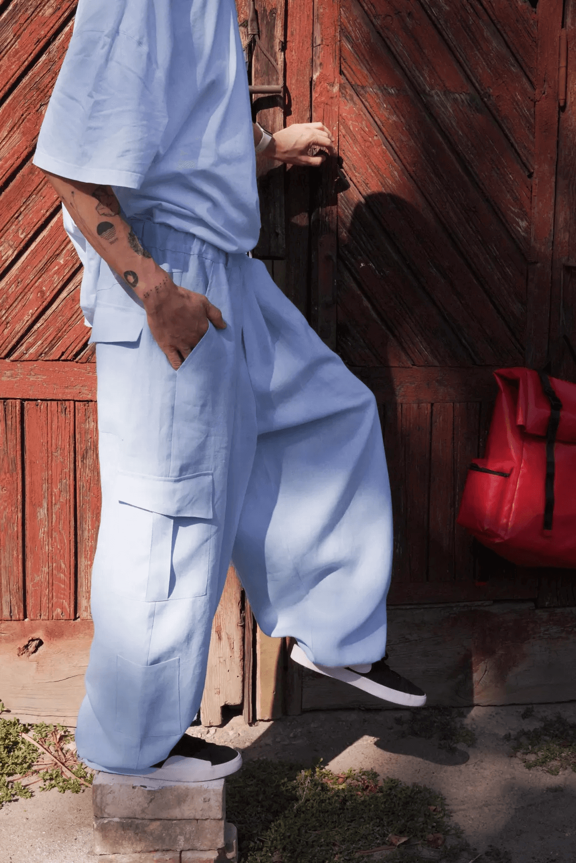 A person standing on a step wearing light grey harem linen pants with side pockets and an elastic waistband, in front of a wooden gate.